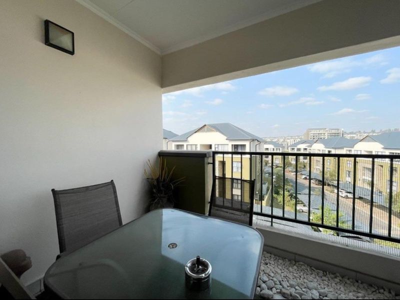 A covered balcony featuring a metal balustrade, pebble flooring, and an exterior wall light, offering an elevated view of the surrounding residential complex.