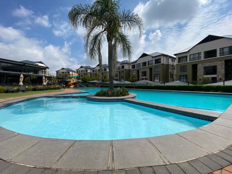 A well-maintained communal swimming pool area featuring paved surrounds, landscaped gardens, and a central palm tree within an apartment complex.