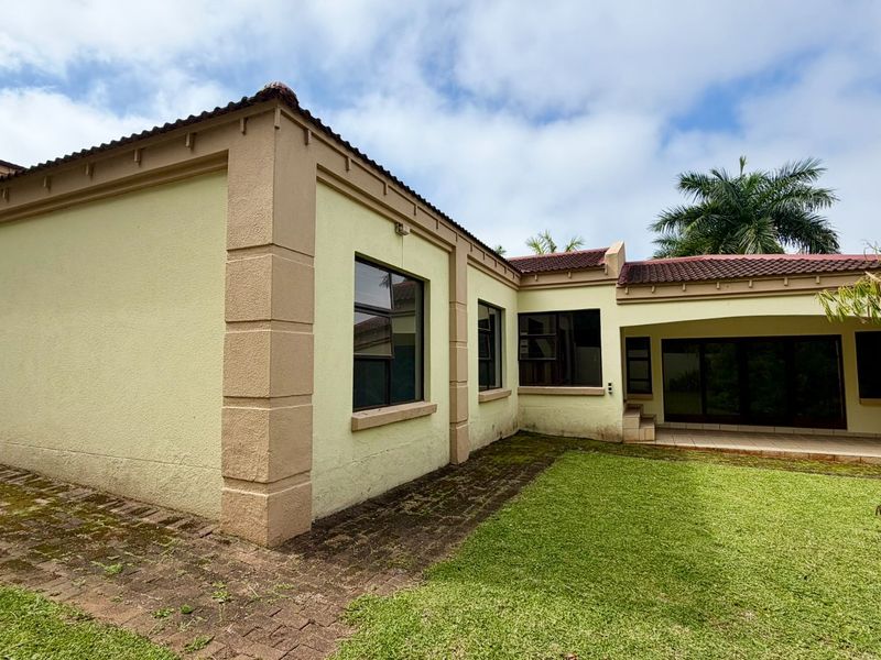 Rear view of the townhouse featuring a covered patio, paved walkway, and established garden area with tiled roof architecture.