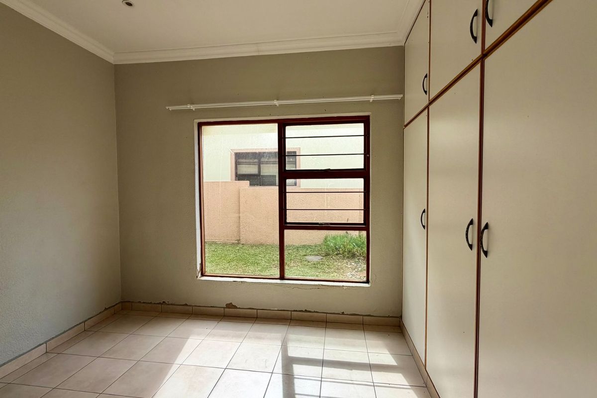 An empty bedroom featuring tiled flooring, built-in cupboards, and a window with security bars, requiring minor cosmetic repairs to the lower wall finishes.