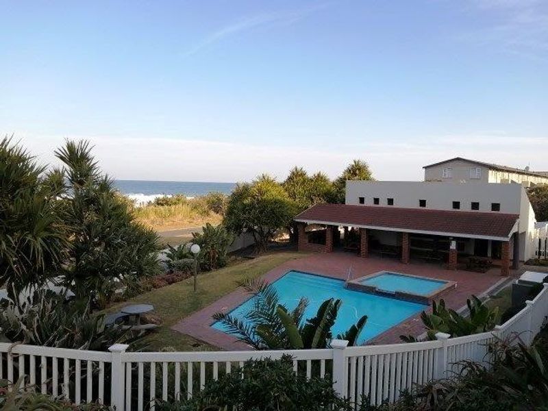 An elevated view of a communal swimming pool area featuring a tiled deck, covered patio, and landscaped garden with ocean views in the distance.
