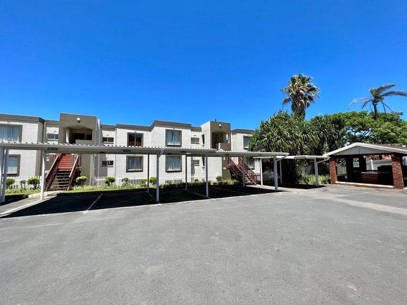 Exterior view of an apartment complex featuring covered parking bays, paved driveway, and a communal brick braai area.