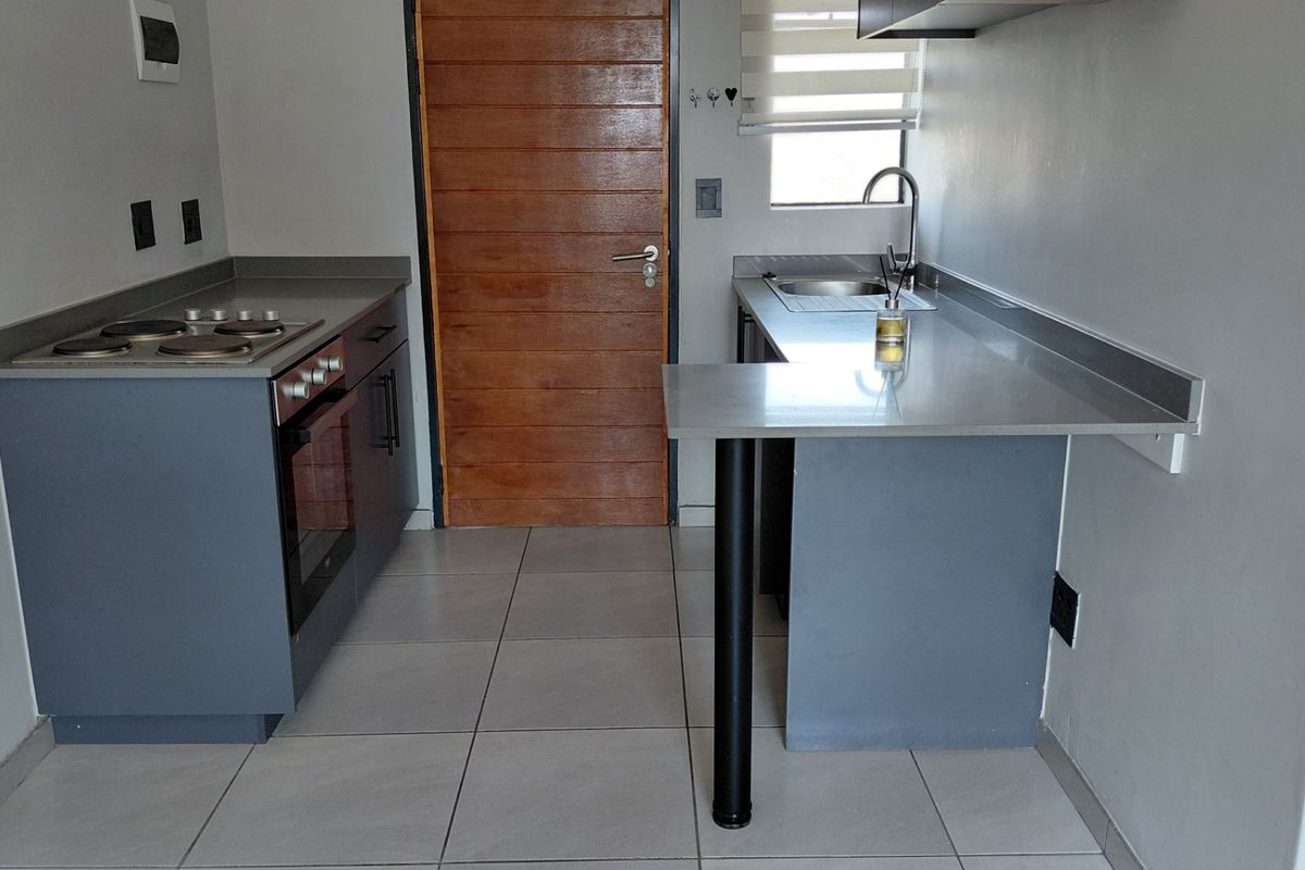 A compact, modern kitchen featuring grey cabinetry, stone countertops, a built-in oven, electric hob, and tiled flooring.