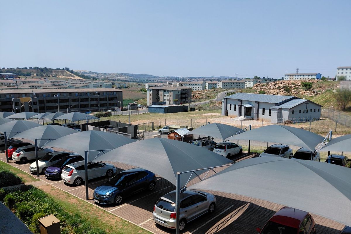 An elevated view of a residential apartment complex parking area featuring multiple shade cloth carports and paved parking bays.