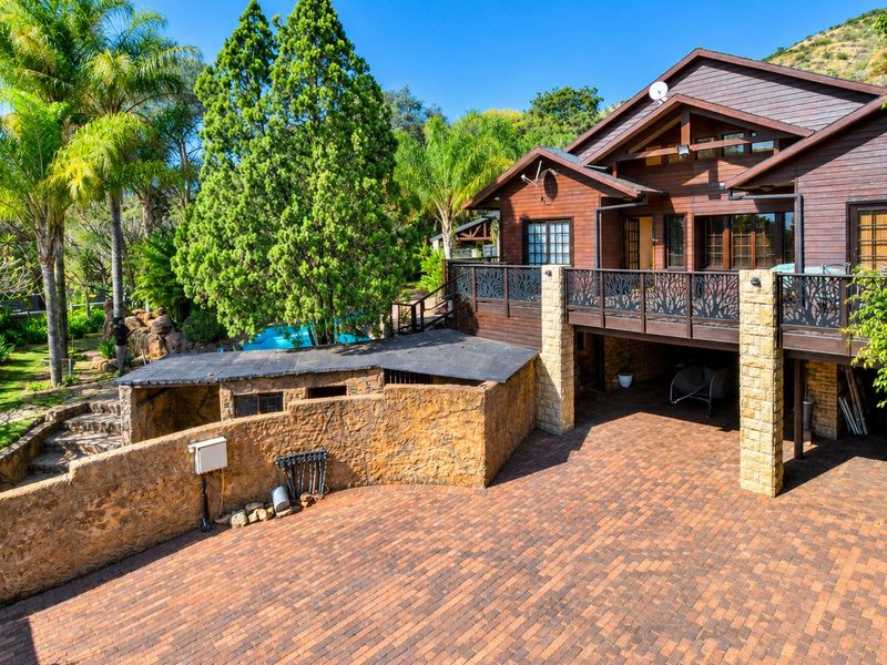 Rear view of a multi-level timber-clad house featuring a raised deck, stone masonry walls, and a paved driveway area set within a landscaped garden.