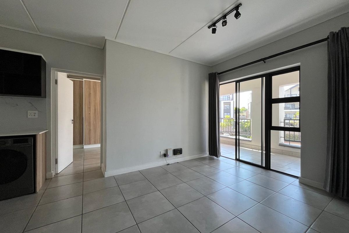 Bright open-plan living area featuring neutral tiled flooring, modern track lighting, and direct access to a private balcony through sliding glass doors.