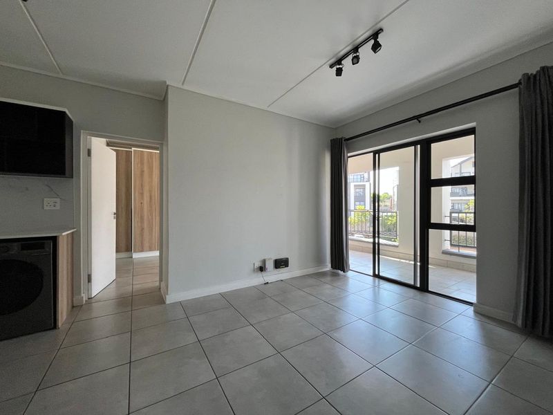 Bright open-plan living area featuring neutral tiled flooring, modern track lighting, and direct access to a private balcony through sliding glass doors.