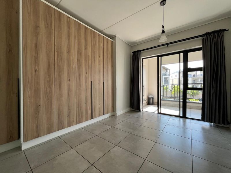 A bright, empty bedroom featuring modern wood-finish built-in cupboards, neutral tiled flooring, and a sliding glass door leading to a private balcony.