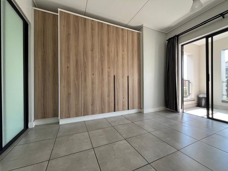 A bright, modern bedroom featuring sleek wood-finish built-in cupboards, large format floor tiles, and direct access to a private balcony through sliding glass doors.