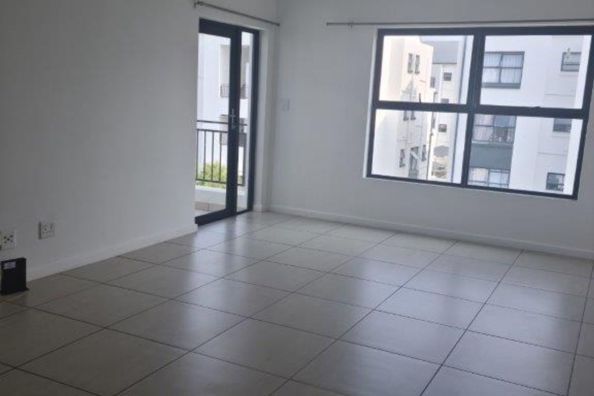 A bright, empty living room featuring neutral tiled flooring, large aluminium-framed windows, and a glass door leading to a balcony.