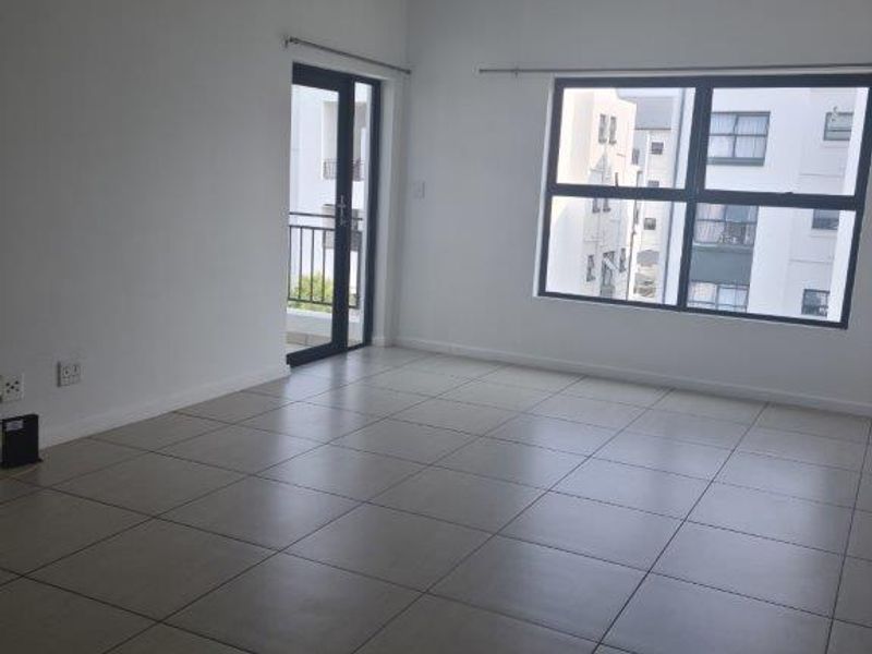 A bright, empty living room featuring neutral tiled flooring, large aluminium-framed windows, and a glass door leading to a balcony.