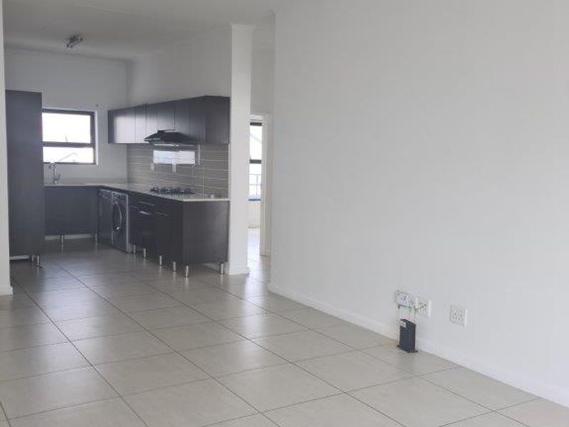 An open-plan living and kitchen area featuring neutral tiled flooring, modern dark cabinetry, and integrated kitchen appliances in a clean, vacant space.