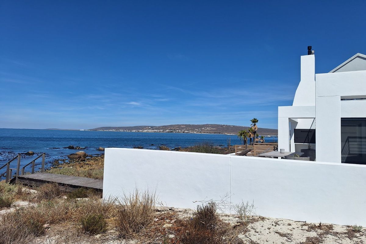 Rear exterior view of a modern white coastal property featuring a chimney, boundary wall, and direct timber boardwalk access to the rocky shoreline.