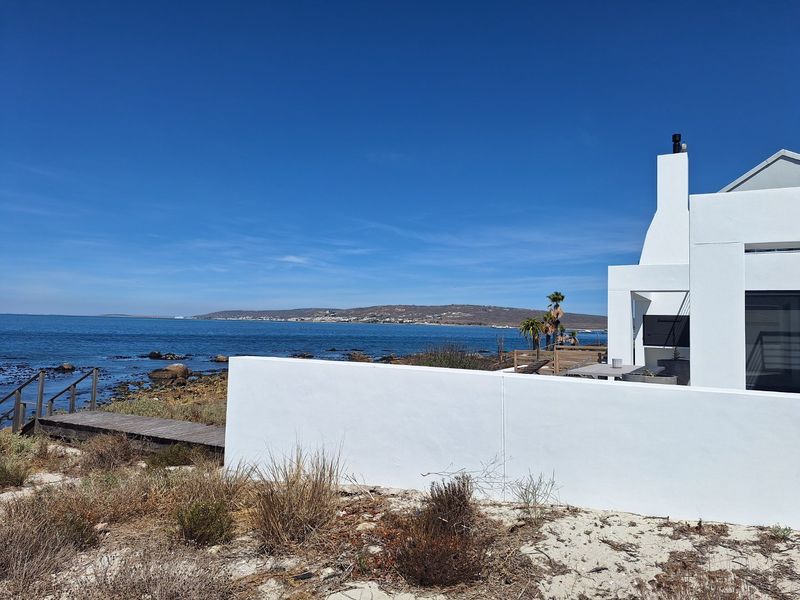 Rear exterior view of a modern white coastal property featuring a chimney, boundary wall, and direct timber boardwalk access to the rocky shoreline.