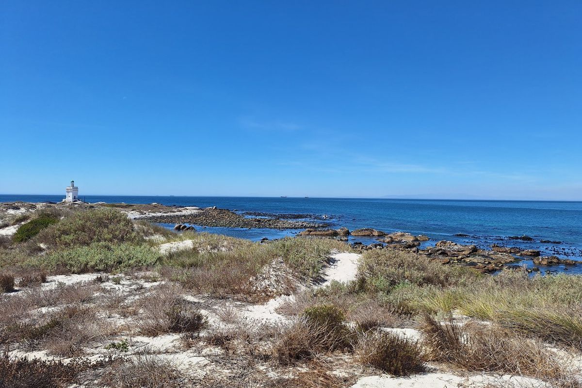 A scenic coastal view featuring sandy dunes, rocky shoreline, and a distant lighthouse overlooking the ocean.