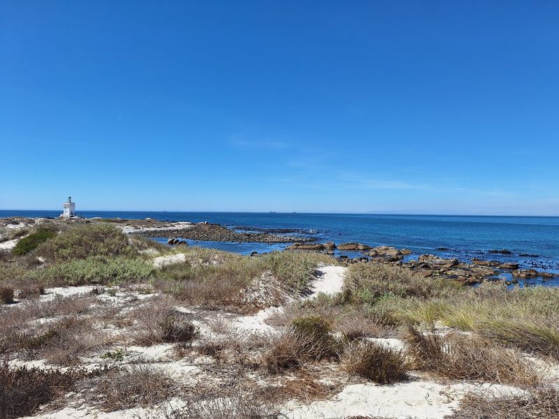 A scenic coastal view featuring sandy dunes, rocky shoreline, and a distant lighthouse overlooking the ocean.