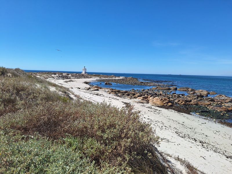 A scenic coastal view featuring a sandy beach, rocky shoreline, and a distant lighthouse under a clear blue sky.