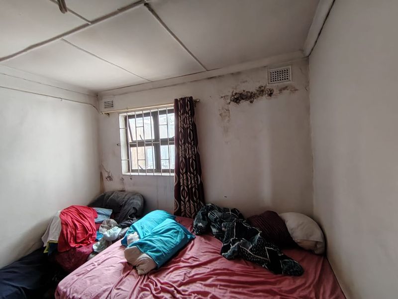 A bedroom featuring security bars on the window and ventilation bricks, currently showing significant signs of wall damp and ceiling deterioration.