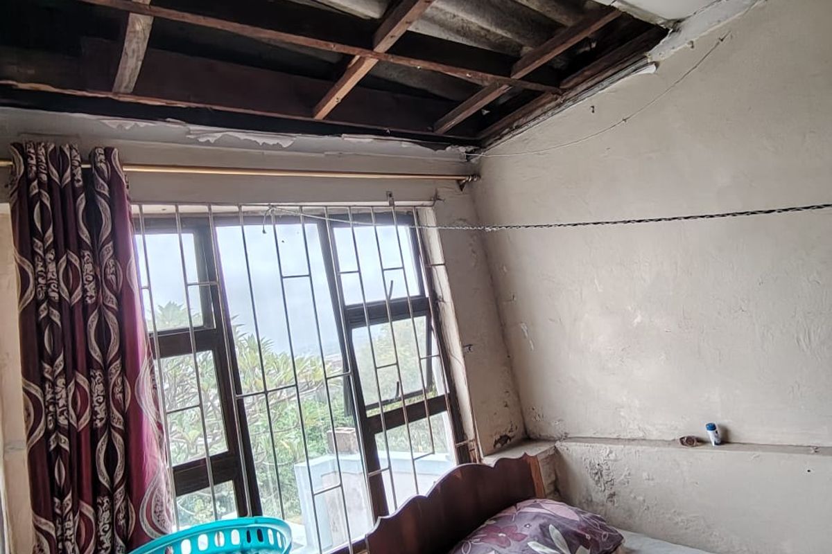 A bedroom featuring security bars on the window and significant ceiling damage with exposed rafters, requiring structural repair and cosmetic renovation.
