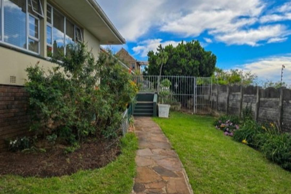 A rear garden view featuring a stone paved walkway leading to a gated entrance, bordered by a concrete wall and established garden beds.