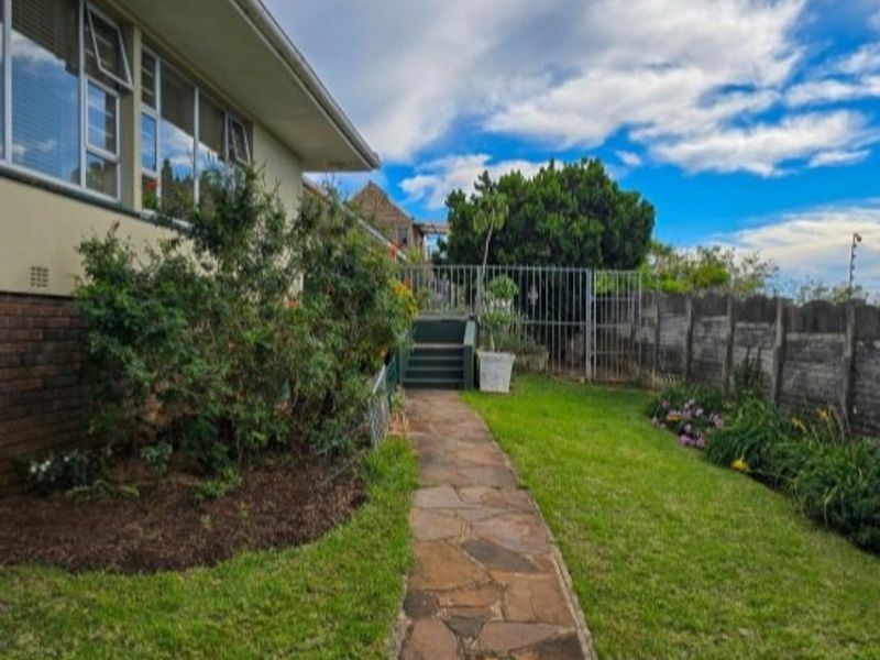 A rear garden view featuring a stone paved walkway leading to a gated entrance, bordered by a concrete wall and established garden beds.