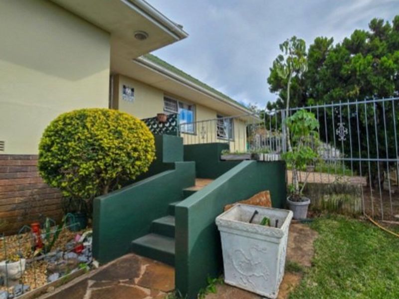 Rear exterior view showing concrete stairs leading to the house, stone paving, and metal security fencing in a garden setting.