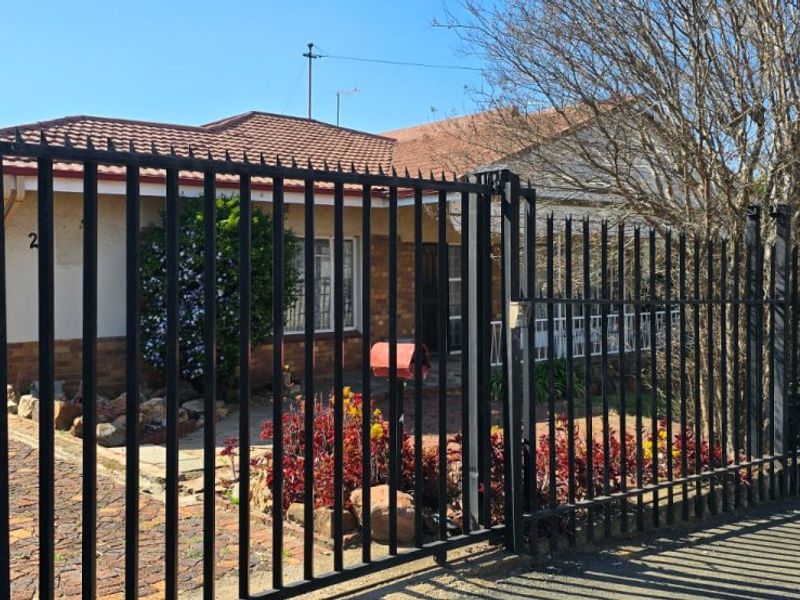 Front view of a residential house featuring a paved driveway, brick facade, tiled roof, and secure metal perimeter fencing.