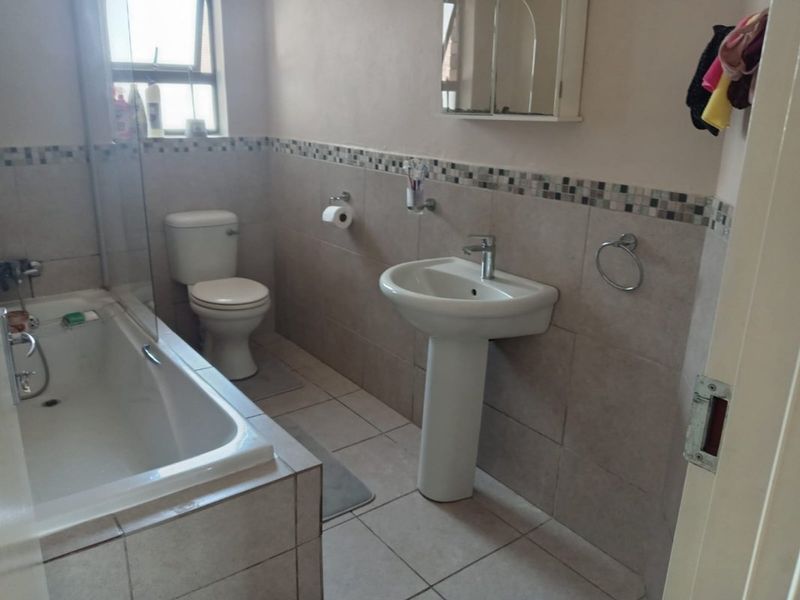 A functional bathroom featuring a pedestal basin, toilet, and a bathtub with a glass shower screen, finished with neutral wall and floor tiles.