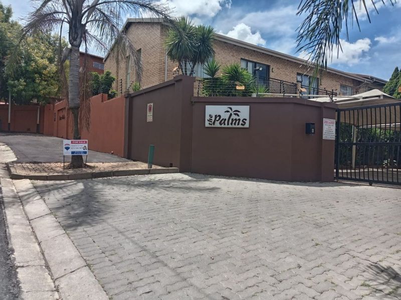 Street-level view of the entrance to The Palms apartment complex, featuring a secure gated access point, brick boundary walls, and paved driveway.