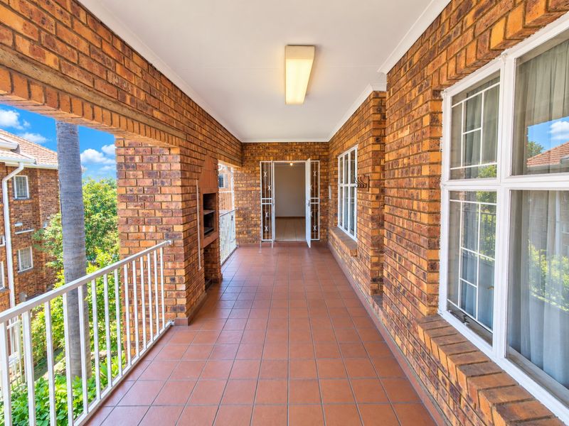Spacious covered balcony featuring exposed brickwork, tiled flooring, a built-in braai, and a metal balustrade, offering a functional outdoor entertainment area.