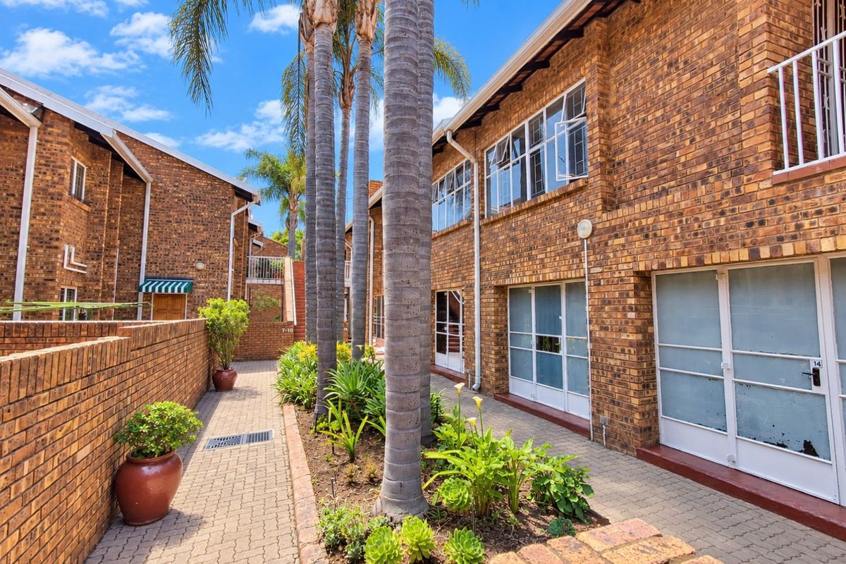 A well-maintained exterior courtyard featuring face brick apartment facades, paved walkways, and established garden beds with mature palm trees.