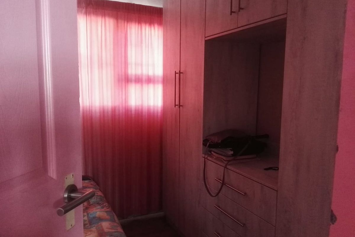 A bedroom featuring built-in wooden wardrobes and laminate flooring, illuminated by natural light through a window with pink curtains.