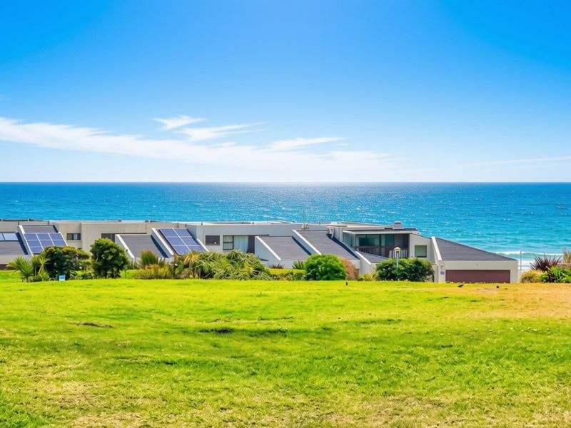 A rear exterior view of a townhouse complex featuring solar panels on the roof, overlooking a grassy lawn and the ocean.