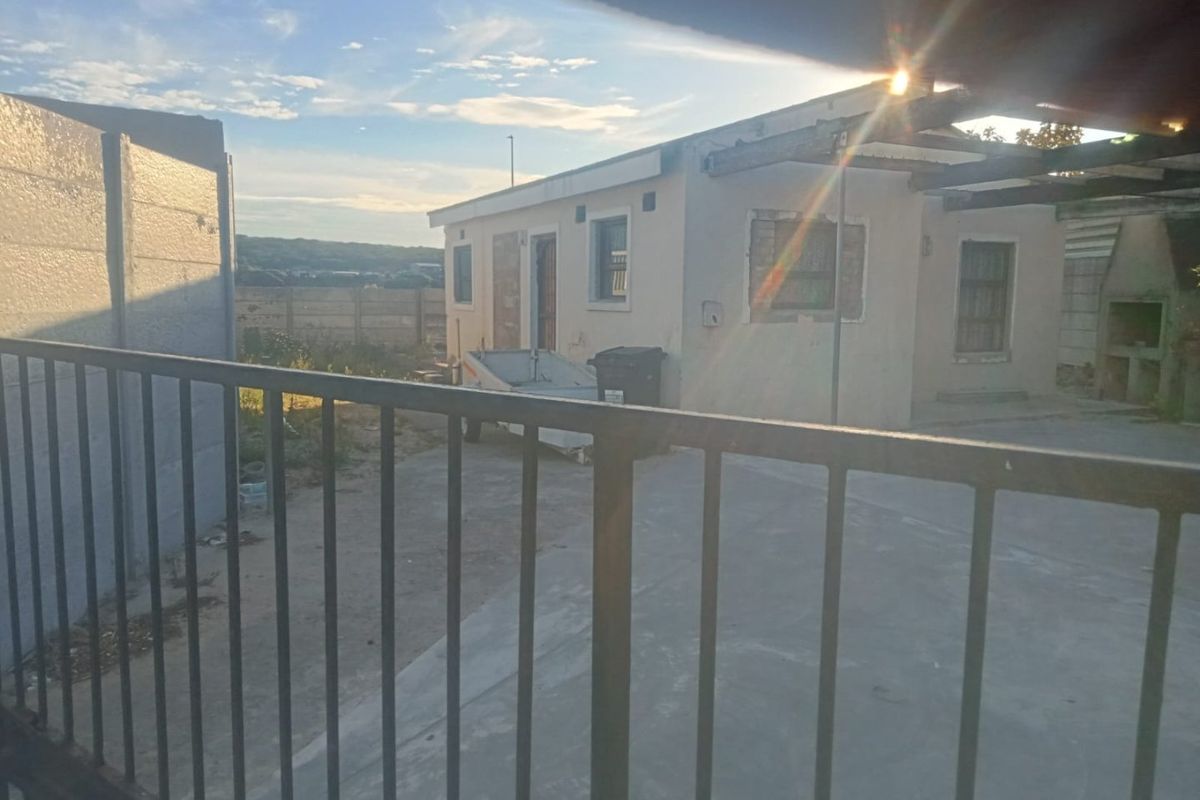 Rear exterior view of the property featuring a paved yard, built-in braai, and perimeter precast walling, partially obscured by a metal security gate.