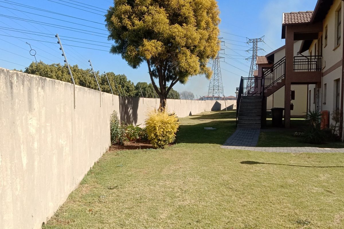 Rear exterior view of the apartment complex featuring a lawn, perimeter wall with electric fencing, and an external staircase leading to upper units.