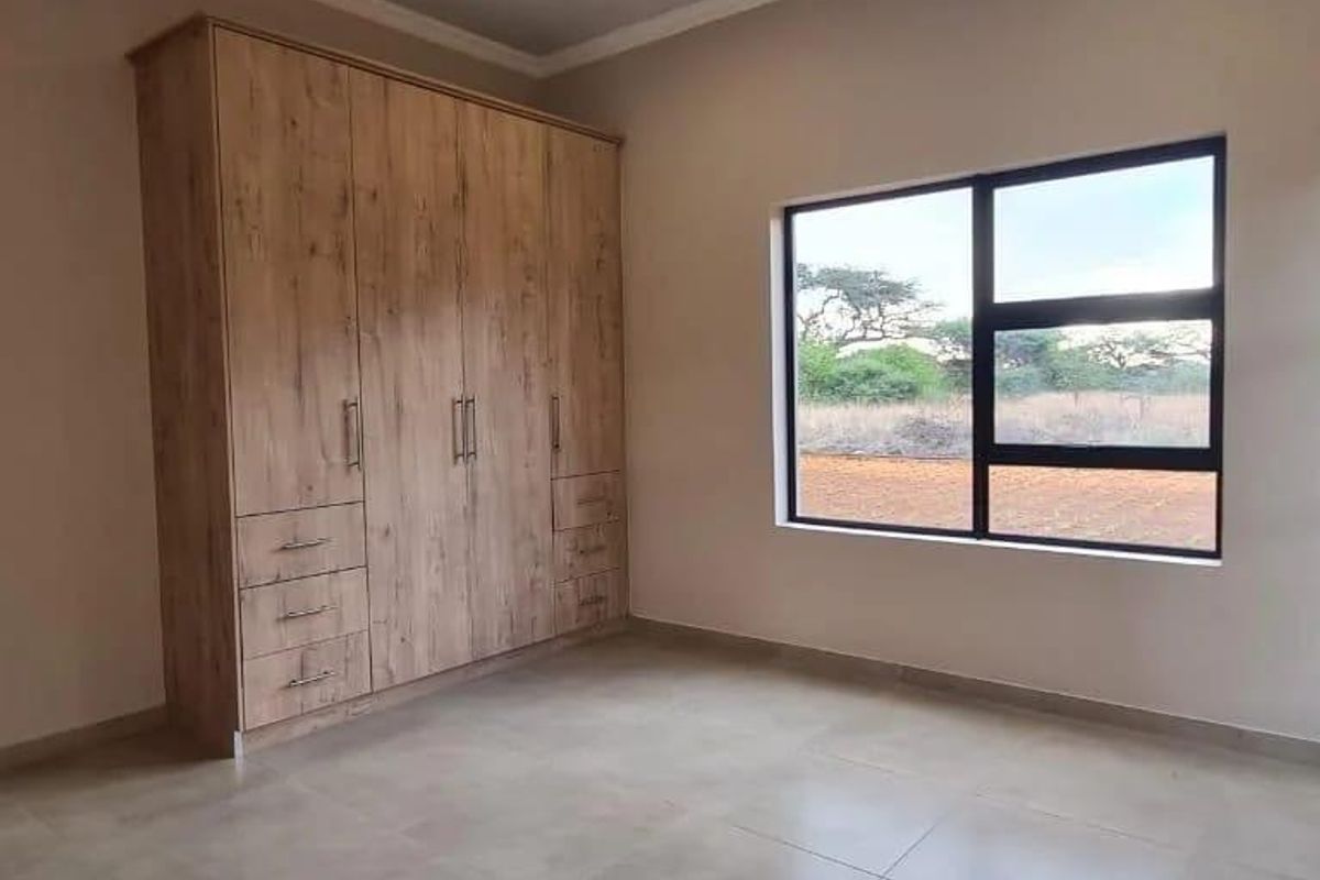 A bright, modern bedroom featuring neutral tiled flooring, built-in wooden cupboards, and large aluminium-framed windows with a natural outlook.