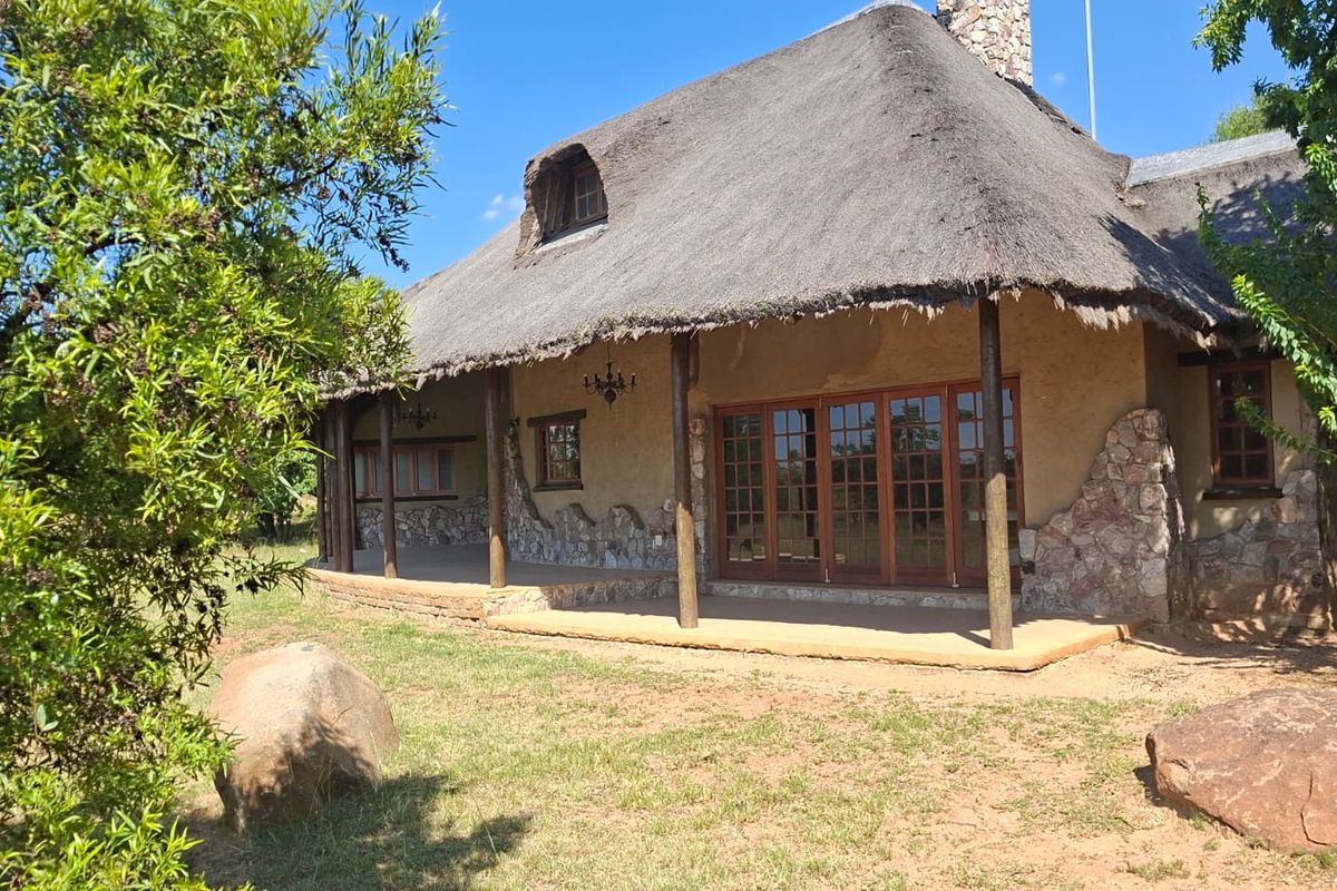 A charming rear exterior view of a residence featuring a traditional thatched roof, stone cladding, and a spacious covered patio with timber-framed glass doors.