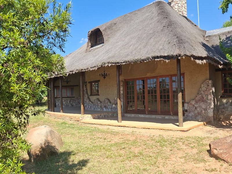 A charming rear exterior view of a residence featuring a traditional thatched roof, stone cladding, and a spacious covered patio with timber-framed glass doors.