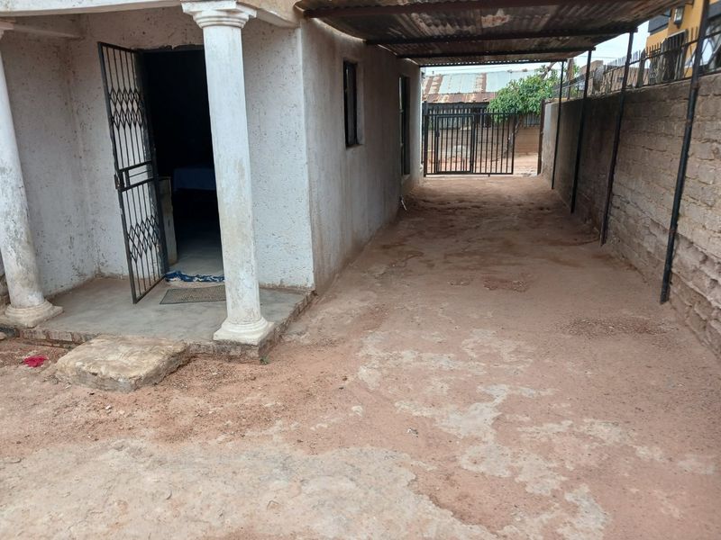 Exterior view of a property featuring a covered carport area with concrete columns, security gate, and boundary wall, showing signs of significant wear.
