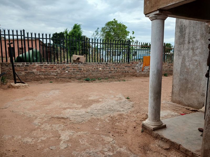 Rear outdoor area featuring a concrete patio column, brick boundary wall, and metal perimeter fencing, requiring landscaping and surface improvements.