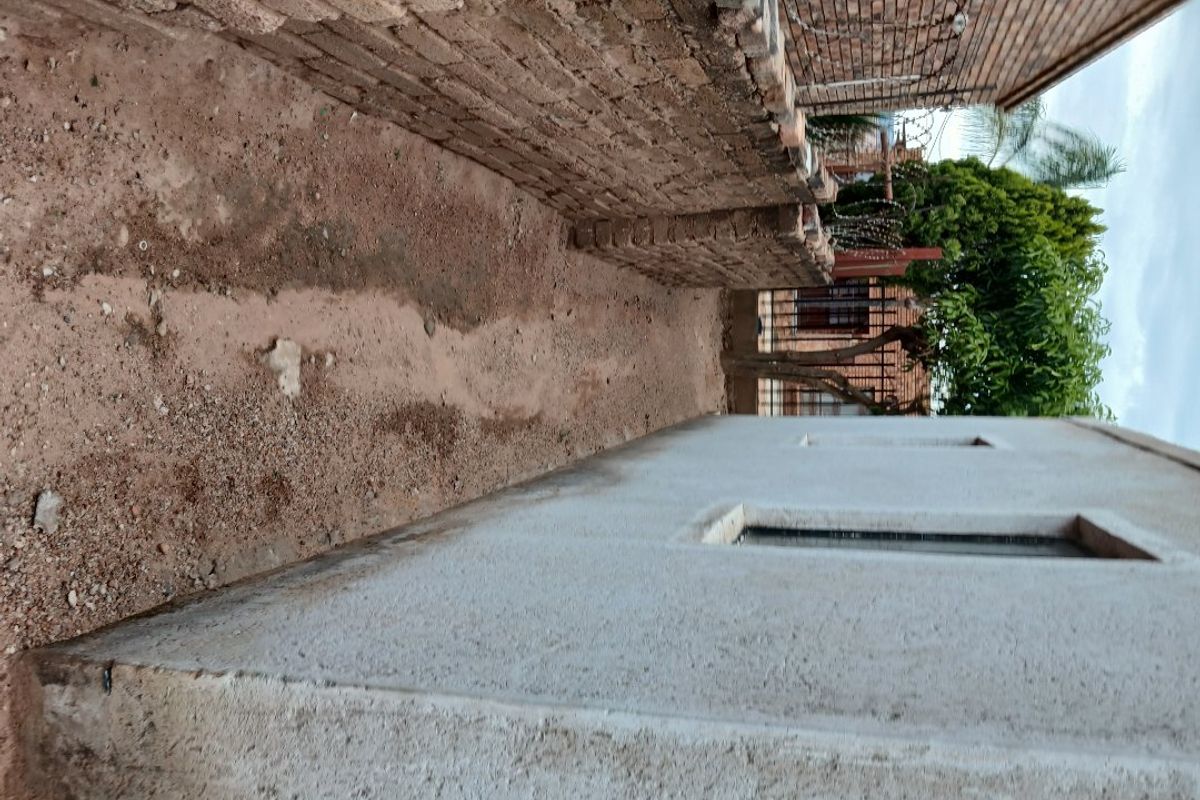 A narrow side passage of the property featuring a plastered exterior wall, brick boundary wall with razor wire, and an unpaved ground surface.