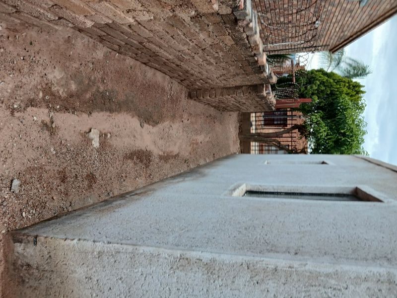 A narrow side passage of the property featuring a plastered exterior wall, brick boundary wall with razor wire, and an unpaved ground surface.
