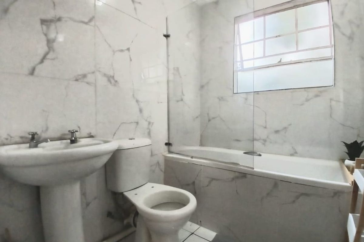 A functional bathroom featuring marble-look wall tiles, a pedestal basin, a toilet, and a bathtub with a glass shower screen.