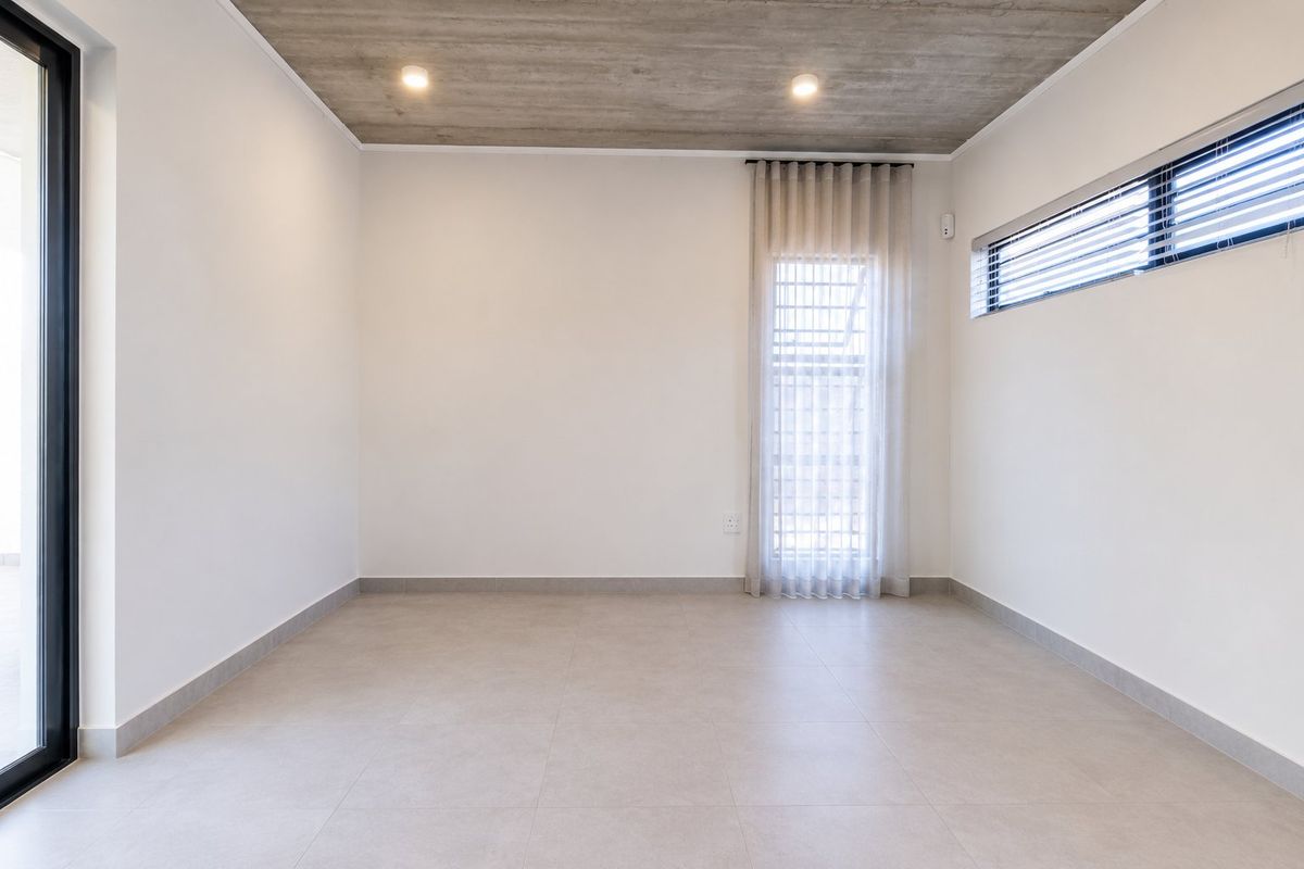 A bright, modern, and empty bedroom featuring an exposed concrete ceiling, neutral tiled flooring, and large glass doors leading to an outdoor area.