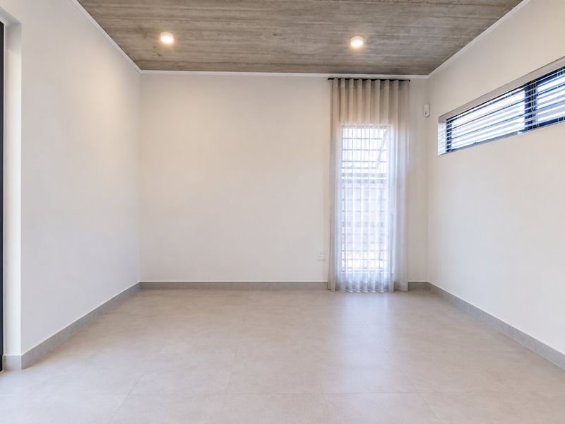 A bright, modern, and empty bedroom featuring an exposed concrete ceiling, neutral tiled flooring, and large glass doors leading to an outdoor area.