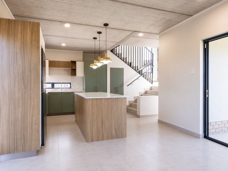 Modern open-plan kitchen featuring a central island, sleek cabinetry, and an exposed concrete ceiling, seamlessly connected to a staircase and outdoor access.