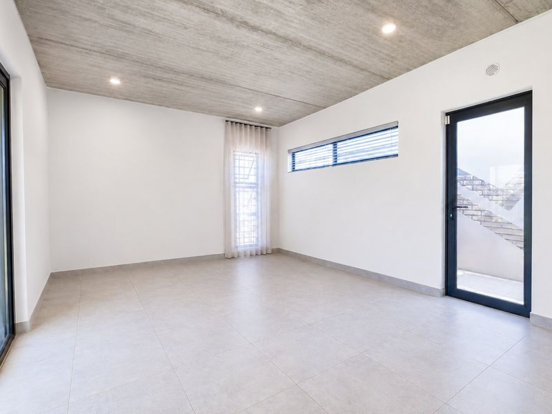 A bright, modern, and empty living space featuring an exposed concrete ceiling, large tiled floors, and direct access to outdoor areas through glass doors.
