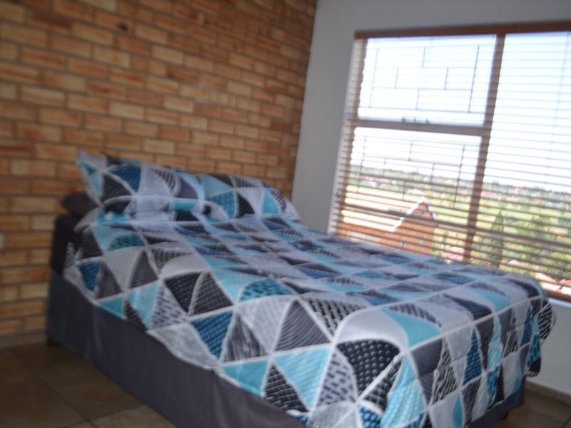 A bedroom featuring an exposed brick wall, tiled flooring, and large windows with venetian blinds, offering natural light and a view.