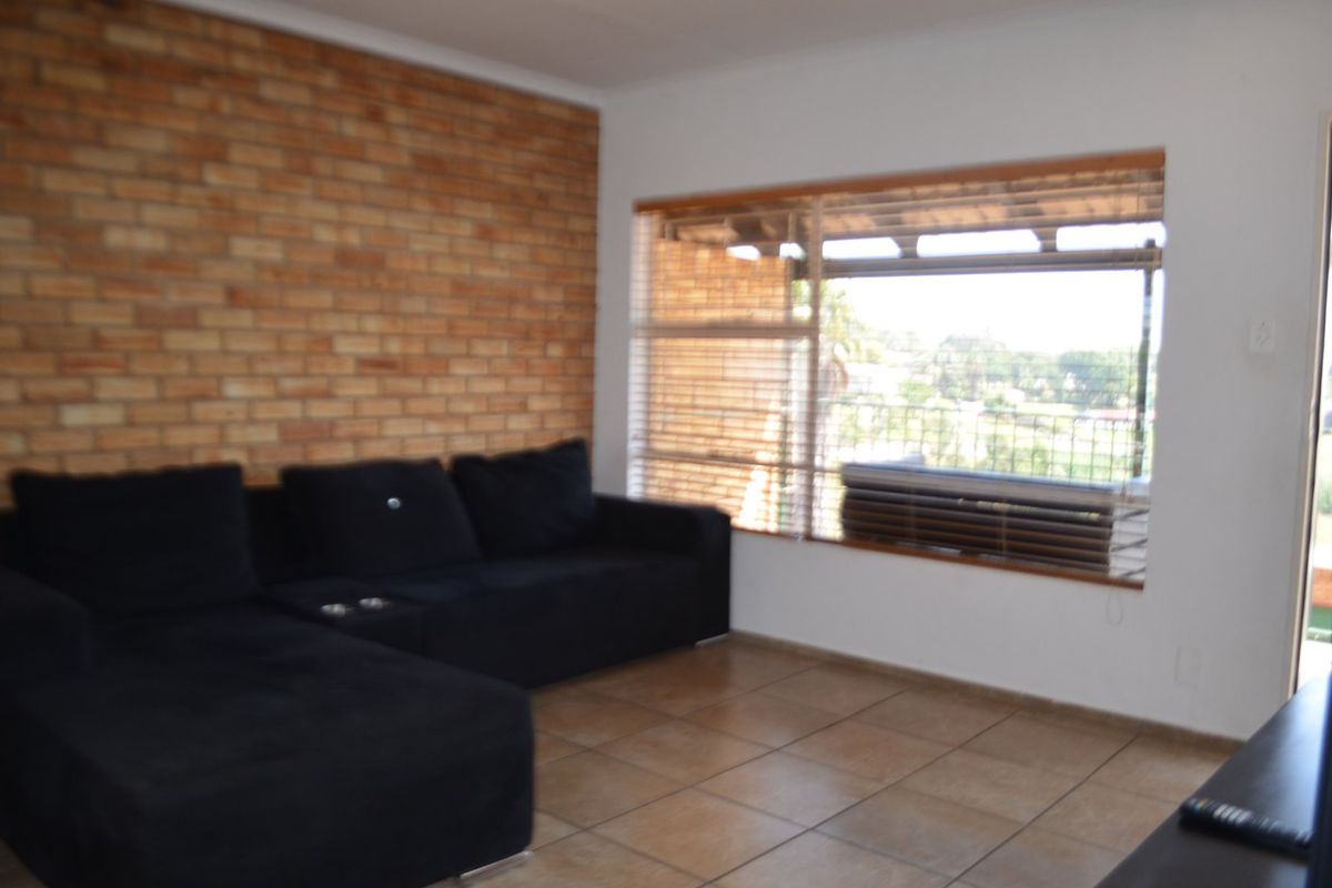 A living room featuring an exposed brick accent wall, tiled flooring, and a large window with venetian blinds providing natural light.