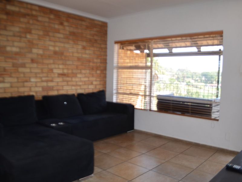 A living room featuring an exposed brick accent wall, tiled flooring, and a large window with venetian blinds providing natural light.